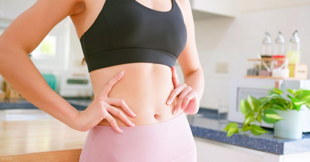 female (model) with toned midsection stands in her kitchen with hands on hips